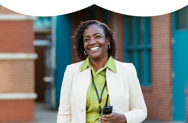 A woman carrying a walkie talkie smiles with campus buildings in the background