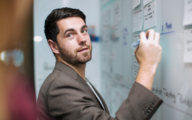 A man in a brown suit stands at a whiteboard holding a marker