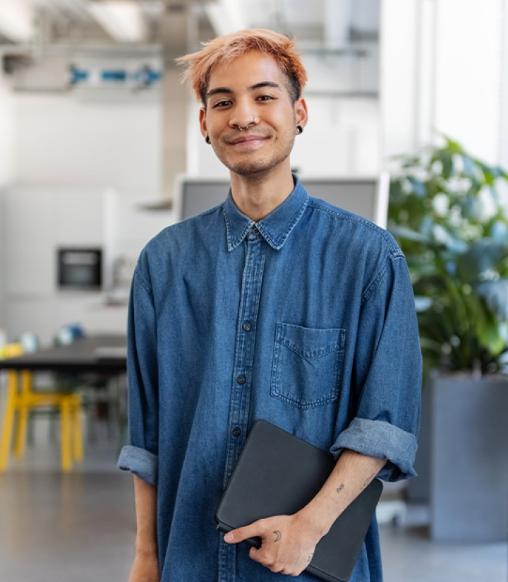 An MEd student stands in an open room with desks and plants  in the background holding a tablet.