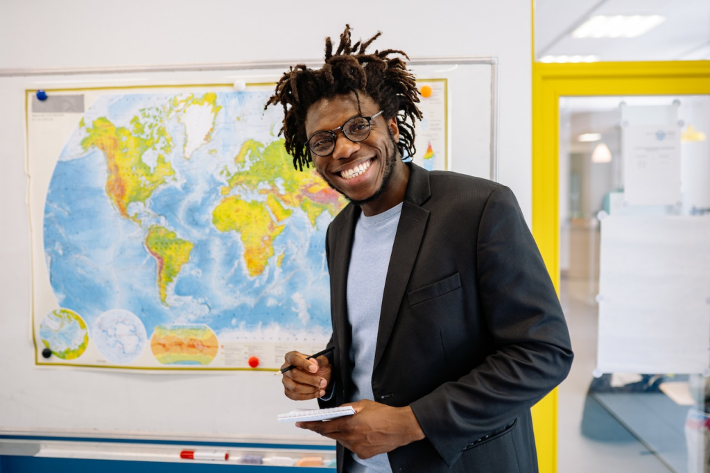 A Black male teacher wearing glasses and a sports coat smiles as he stands before a world map holding a pen and pad.