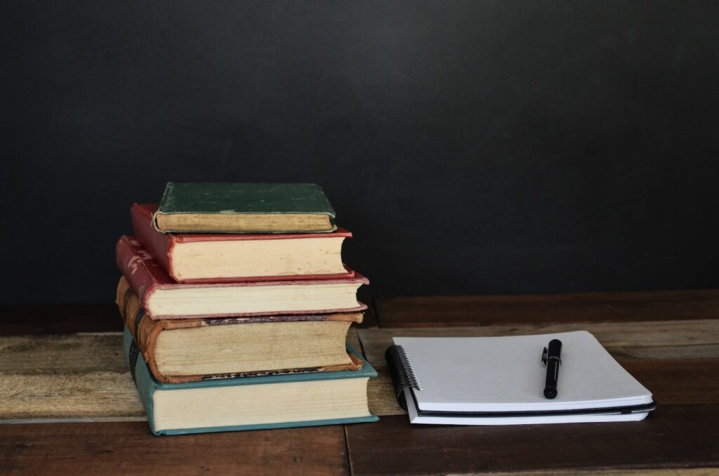 A stack of books sit next to a notebook with a pen.