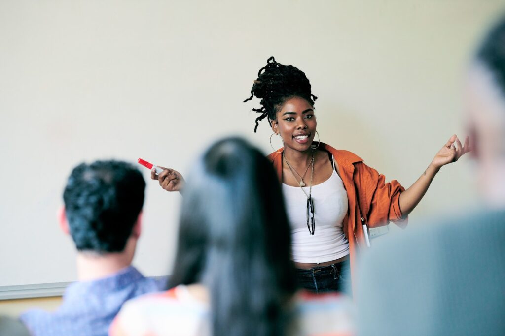 A smiling Black female instructor standing in front of a whiteboard gestures emphatically.