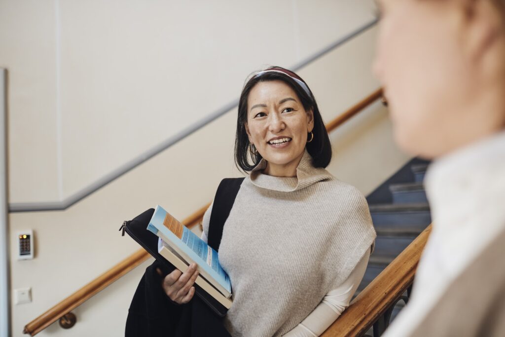 A smiling female Asian teacher carrying books encounters a female student on a school stairwell.