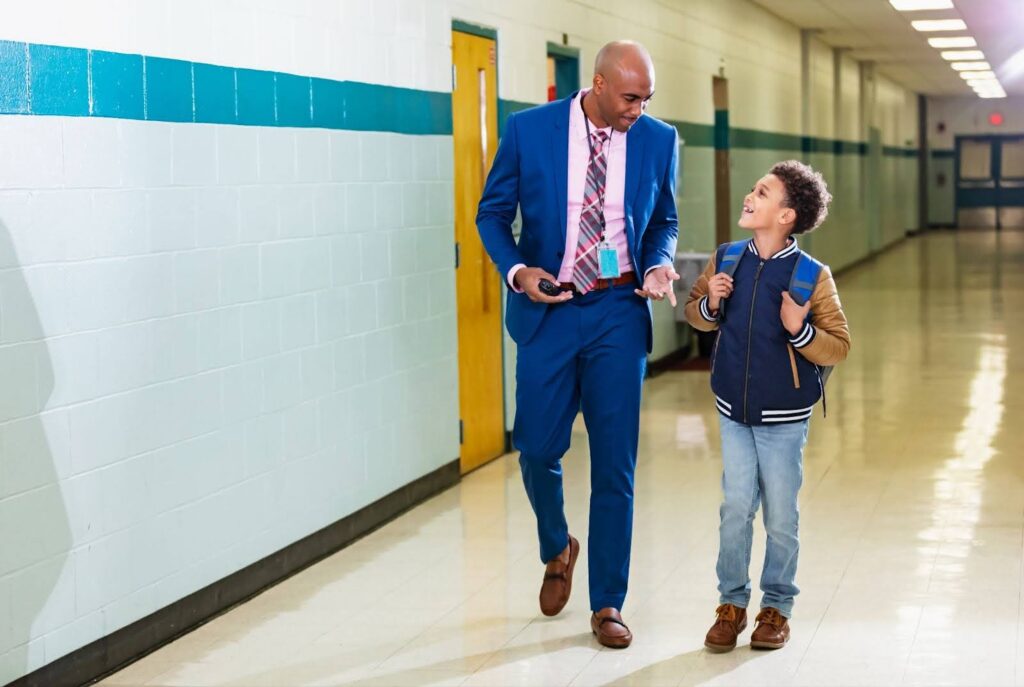 A school principal walks down a school hallway conversing with a middle school student.