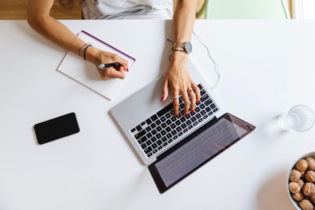 Overhead shot of a laptop computer on a table; the user is typing with her left hand and taking notes on a notepad with her right hand.