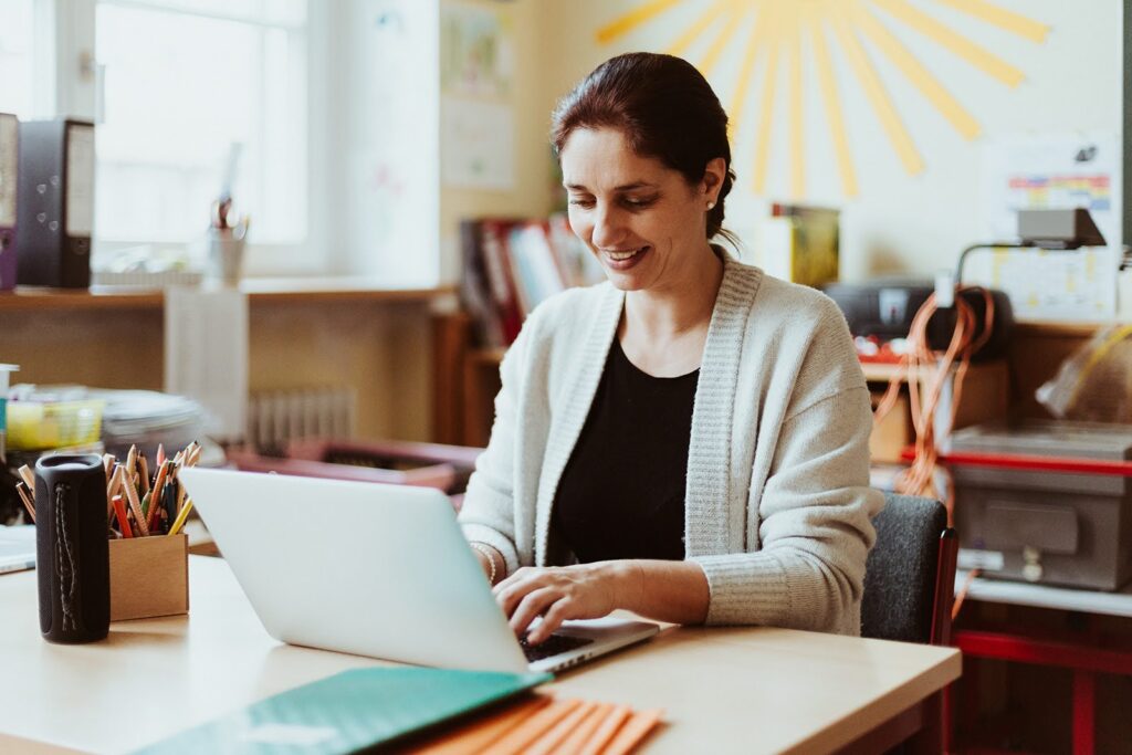 A smiling teacher sits at her desk, typing at a laptop computer.