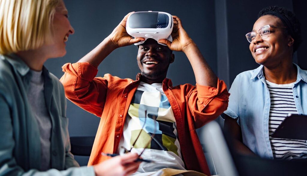 Three young adult students smile and laugh as one prepares to use a virtual reality headset.