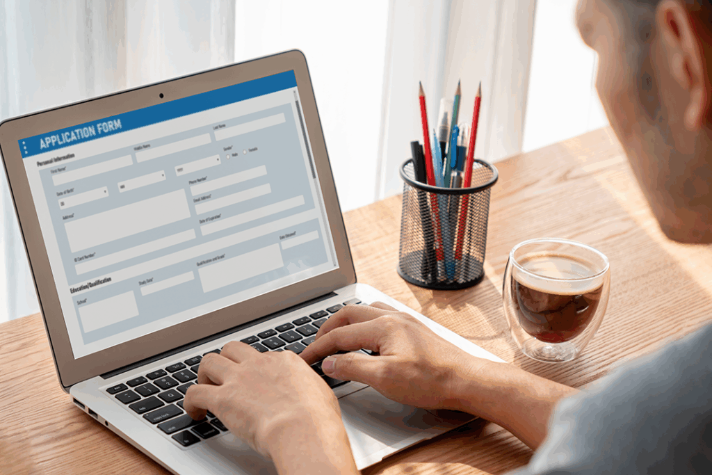 A person uses a laptop to fill out an online application form; a cup of coffee and a container of pencils and pens sit beside the computer.