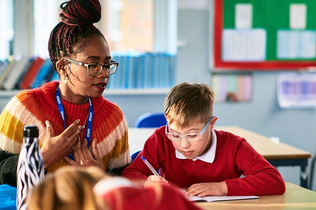 Teacher wearing a striped sweater helps a young student with glasses during class.