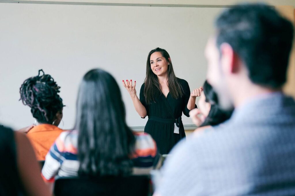 A woman stands at the front of a classroom, smiling and gesturing as she speaks to a seated group of adults.